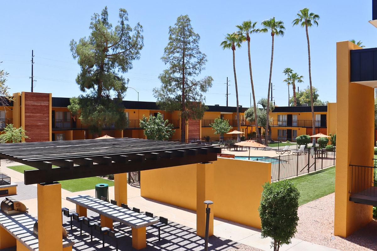 A view of a sunny outdoor area at a hotel or apartment complex. There are palm trees, a well-maintained grassy lawn, a swimming pool surrounded by orange umbrellas, and outdoor seating under a wooden pergola. The buildings are painted in vibrant colors, contributing to a relaxed, tropical atmosphere.