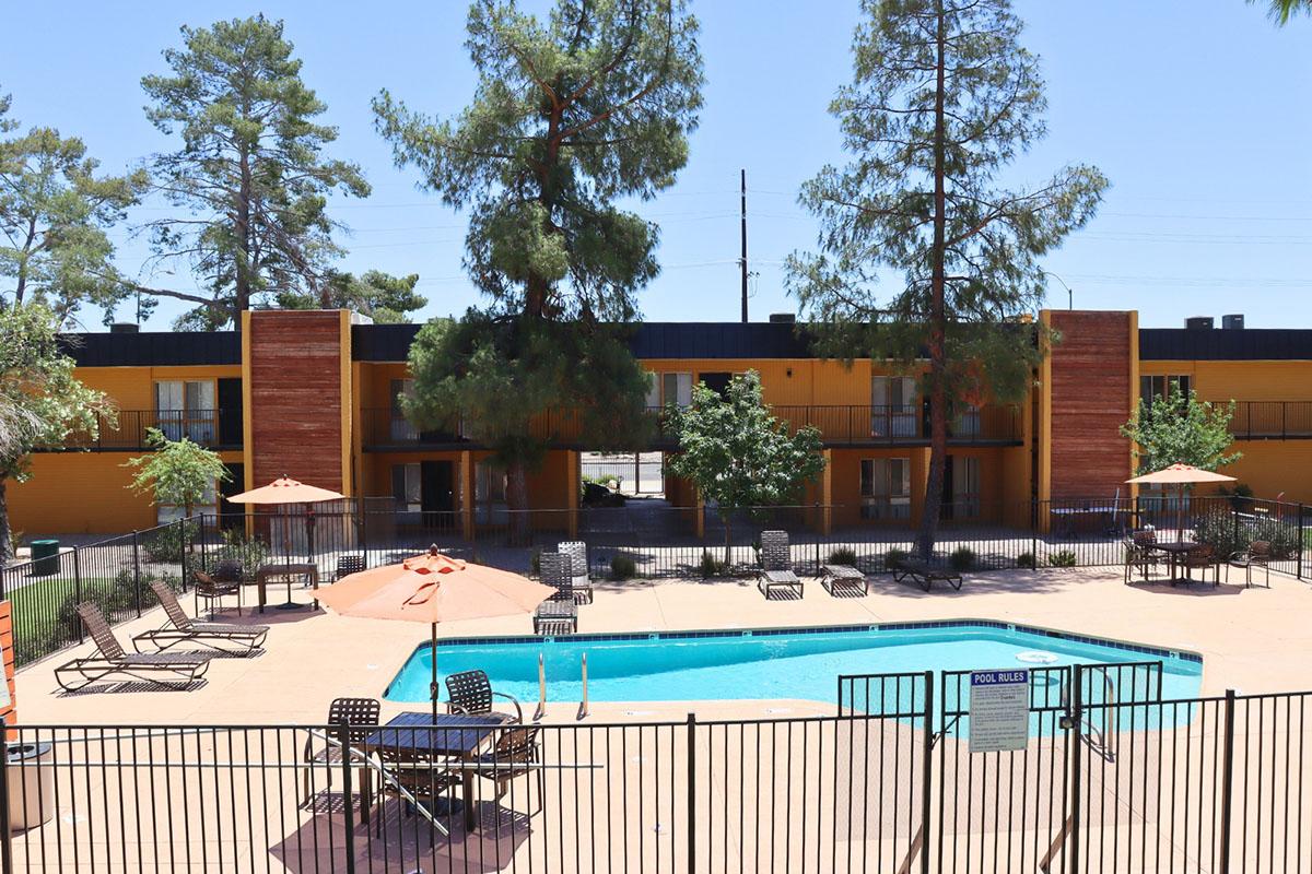 View of a swimming pool surrounded by lounge chairs and umbrellas, set within a fenced area. In the background, there are two-story buildings painted in warm colors and tall trees providing shade. The sky is clear and blue.
