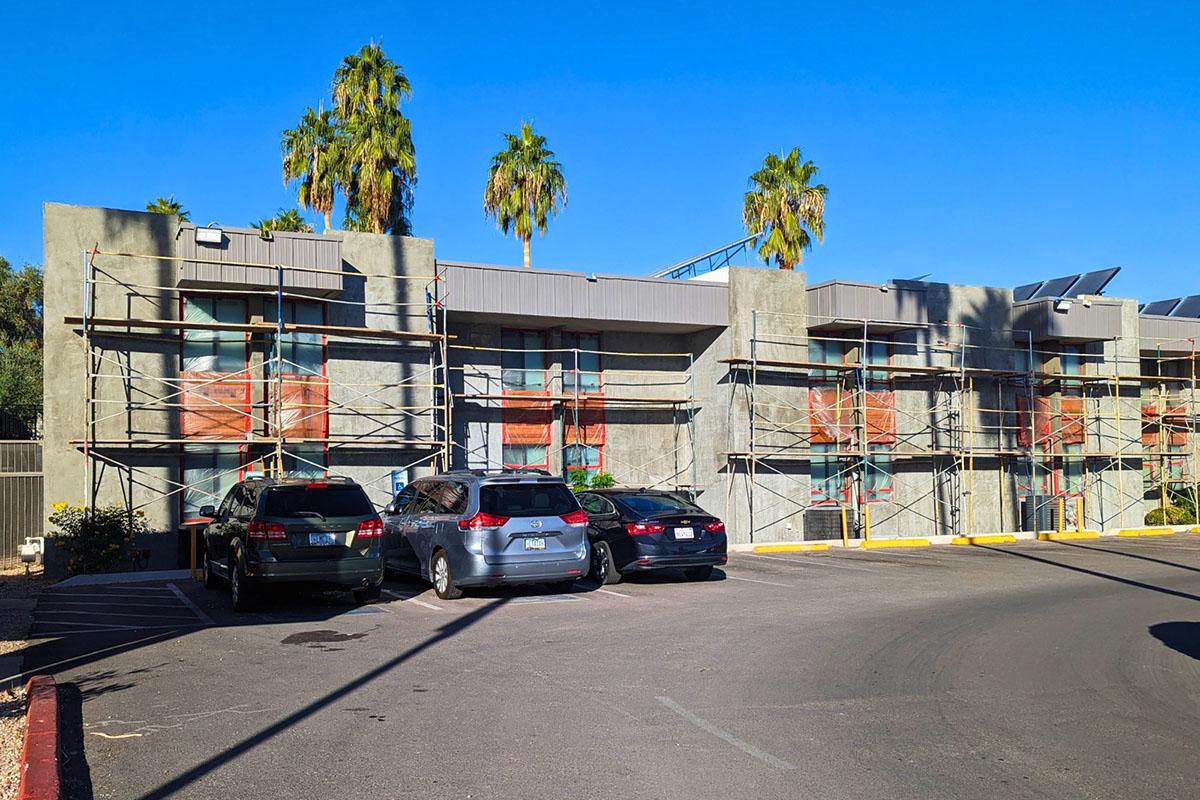 A modern building under renovation with scaffolding on the exterior. The structure features multiple windows and palm trees in the background. Several parked cars are visible in front of the building, and the sky is clear and blue, indicating a sunny day.