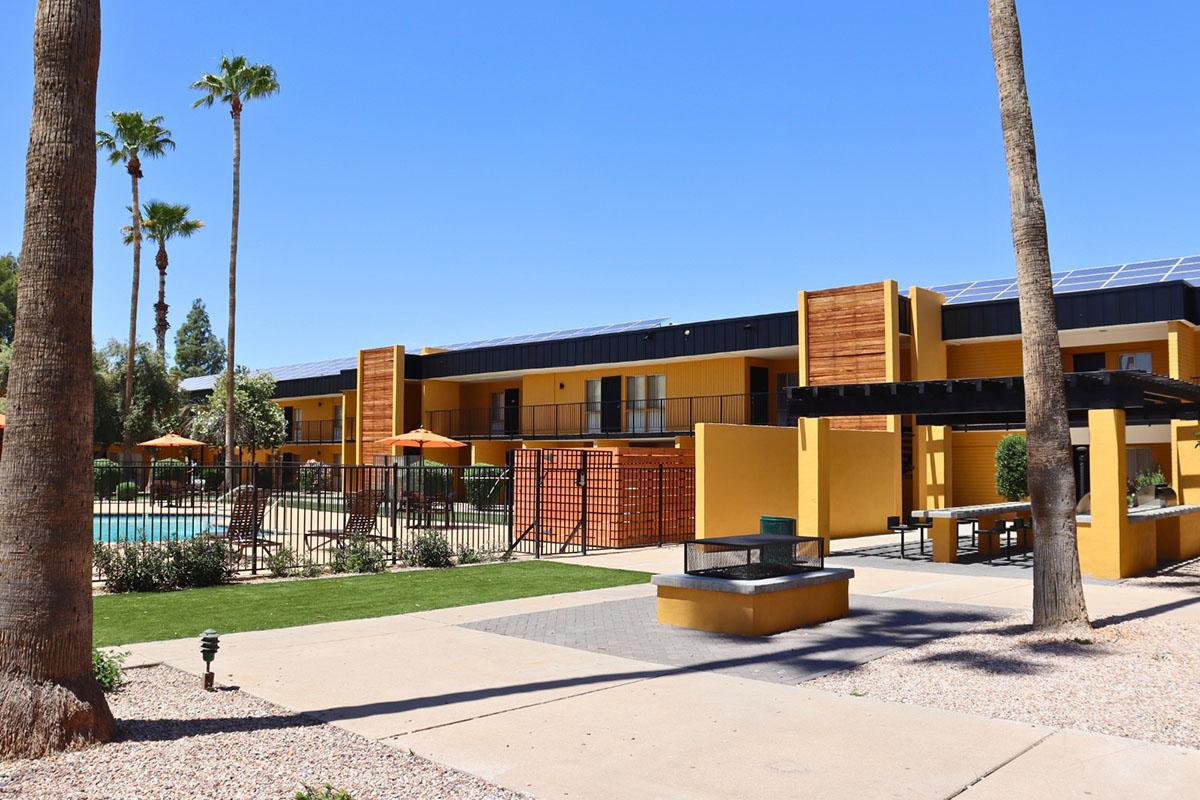 A vibrant courtyard area of an apartment complex featuring a swimming pool, palm trees, and orange patio umbrellas. The modern building has solar panels on the roof and is surrounded by decorative fencing and landscaping. Clear blue skies enhance the inviting atmosphere.