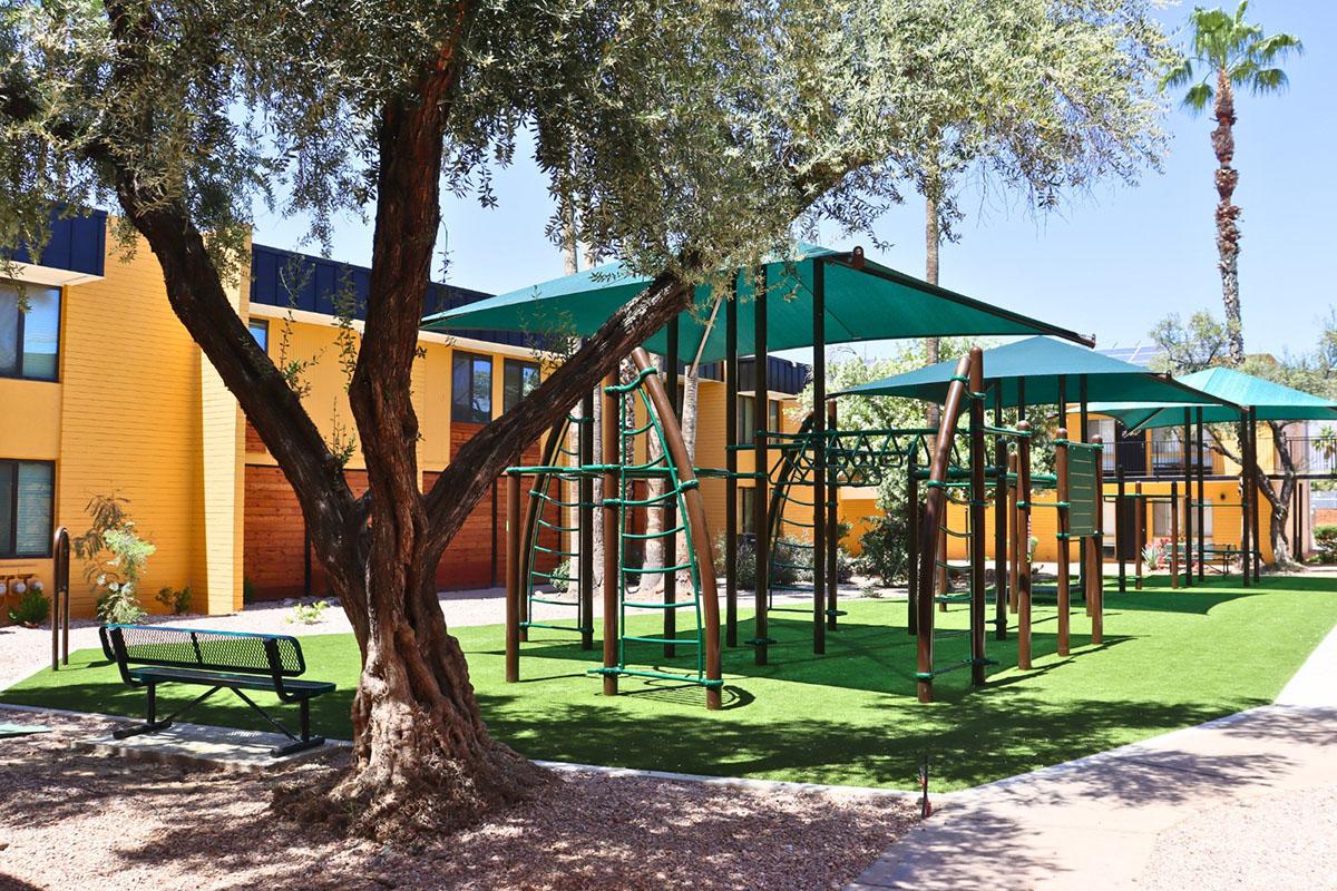 A playground area featuring climbing structures and shaded canopies, surrounded by green synthetic grass and a tree. There is a bench nearby, and the background includes colorful buildings and palm trees, indicating a pleasant outdoor environment.