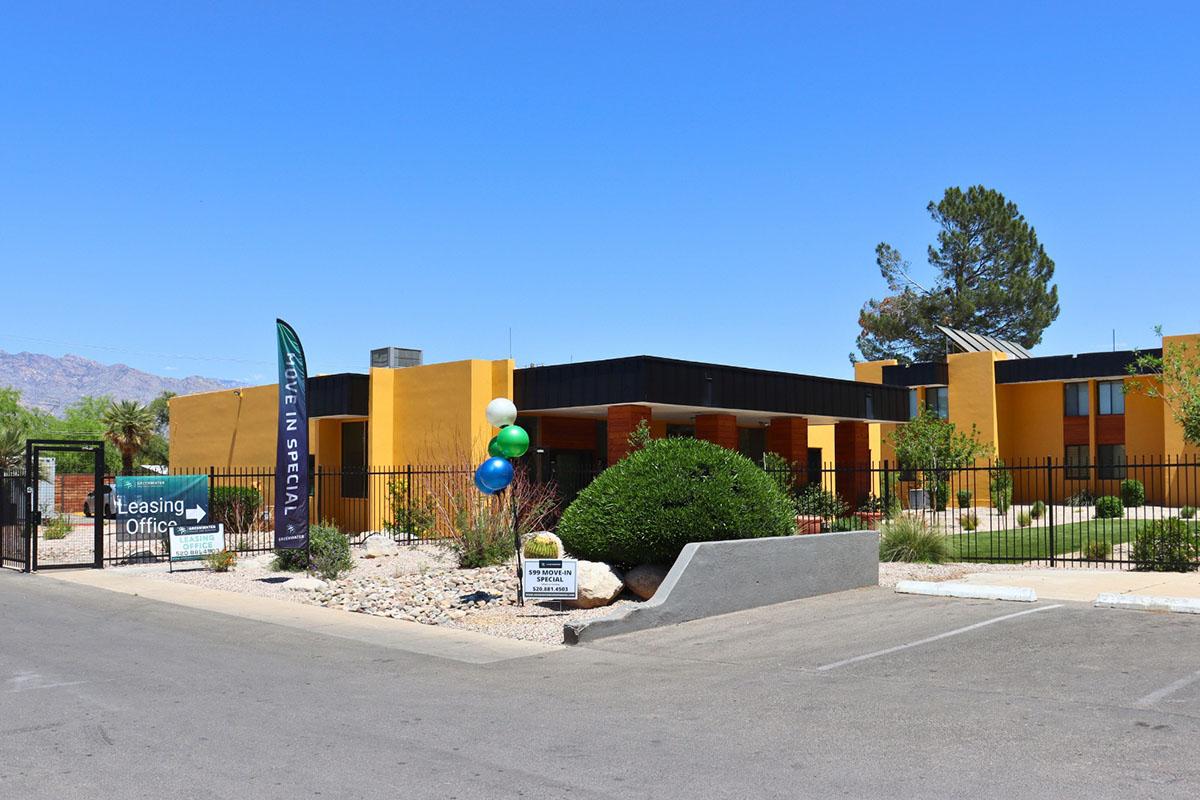 A leasing office for an apartment complex, featuring a gated entrance and colorful balloons. The building has a modern design with an orange and black exterior, surrounded by desert landscaping and decorative rocks. A bright blue sky is visible above.