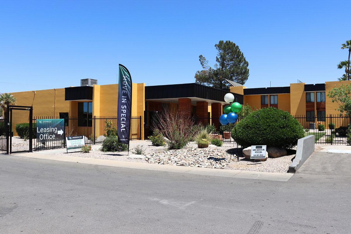 A leasing office building with a colorful balloon display and banners. The exterior features a modern design with desert landscaping, including shrubs and rocks. The scene is set under a clear blue sky, indicating a sunny day. Signs promoting leasing specials are visible in the foreground.