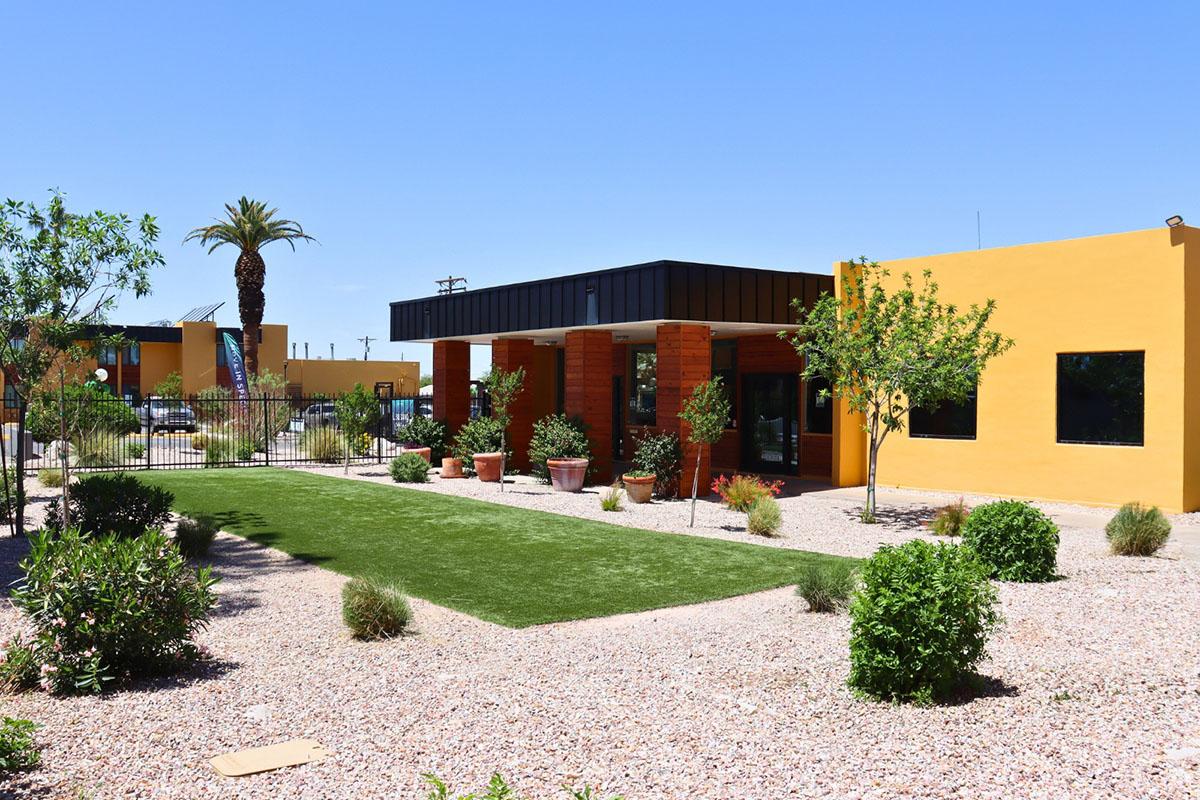 Modern building with a flat roof, featuring large windows and bright orange walls. Surrounded by a landscaped garden with sparse plants, grasses, and palm trees, the area has a clean, contemporary aesthetic under a clear blue sky. A small green lawn complements the design.