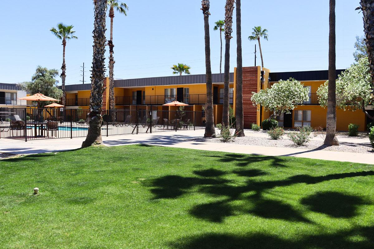 A sunny outdoor view of a hotel complex featuring a swimming pool surrounded by palm trees and orange umbrellas. The building has a yellow exterior with balconies, set against a blue sky. Lush green grass covers the foreground, enhancing the vibrant atmosphere.