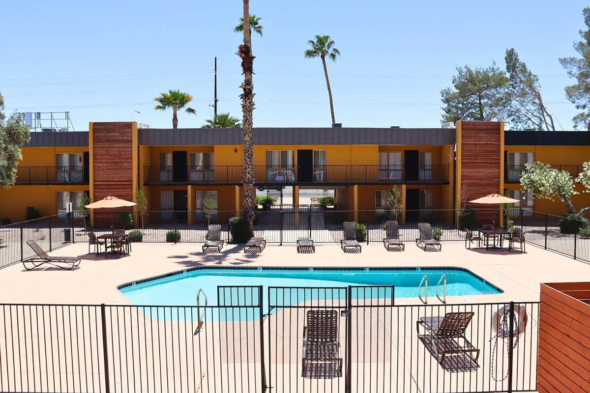 A view of a modern hotel with a swimming pool surrounded by lounge chairs and umbrellas. Yellow and brown building exteriors with palm trees in the background. The area is well-lit and inviting, showcasing a sunny day. The pool area is enclosed with a fence for safety.