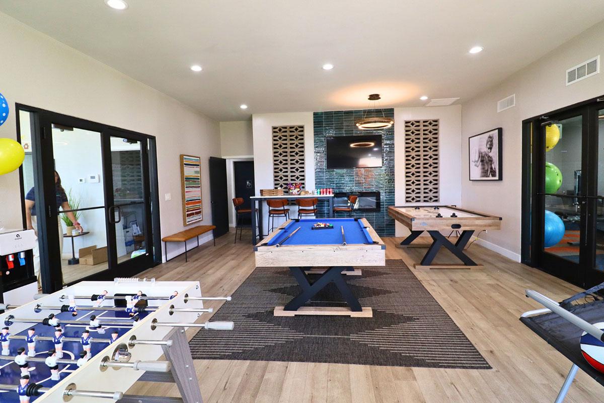 A bright, modern game room featuring a blue pool table and a foosball table. The space includes large windows, stylish decor, and a comfortable seating area. Colorful balloons add a festive touch, while a wall with patterned tiles and a bar area contribute to the inviting atmosphere.