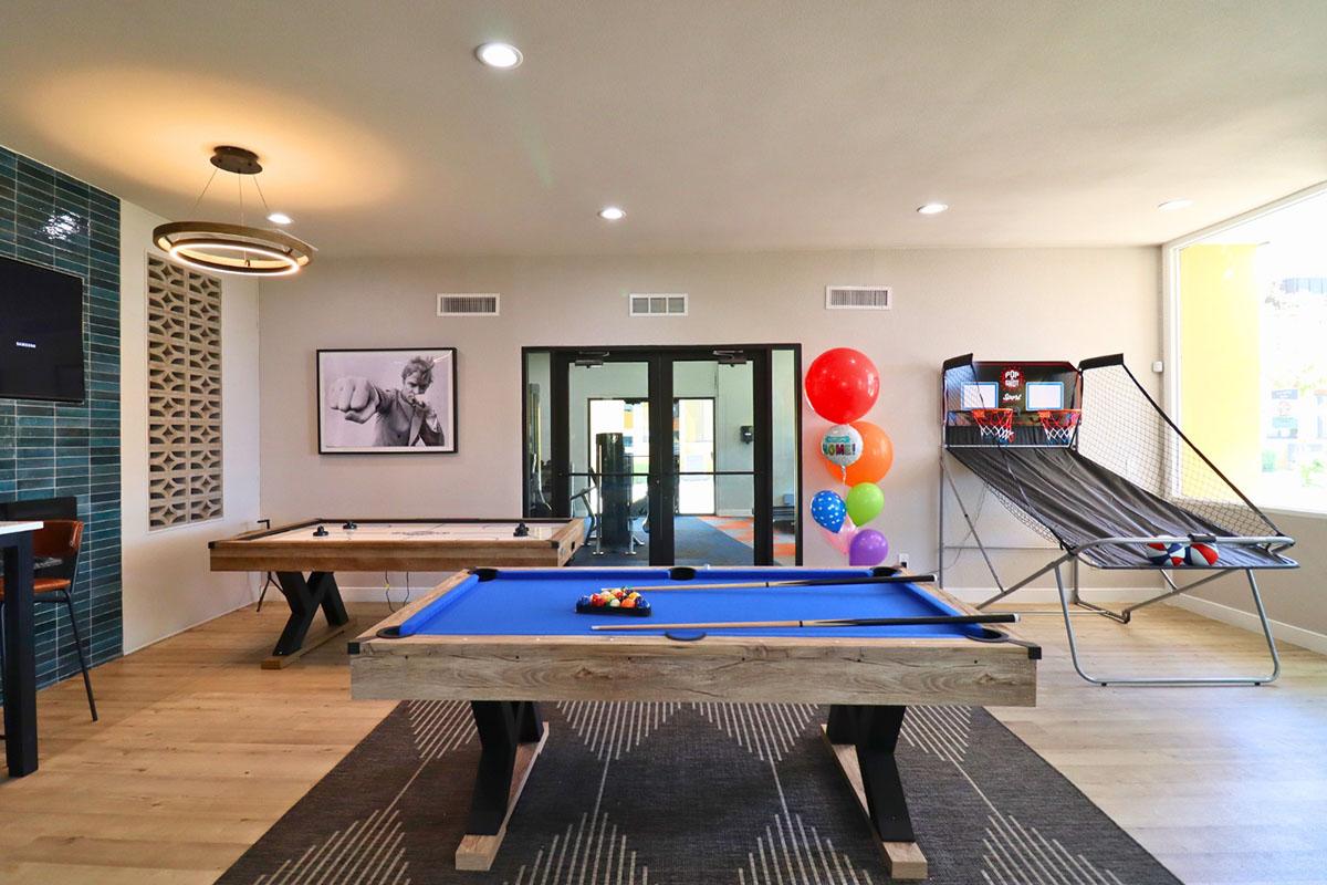 A modern recreational room featuring a blue pool table, a foosball table, a large wall-mounted television, and colorful balloons. The space has bright lighting and contemporary decor, with a view of glass doors leading to another area. A playful atmosphere, perfect for socializing and entertainment.