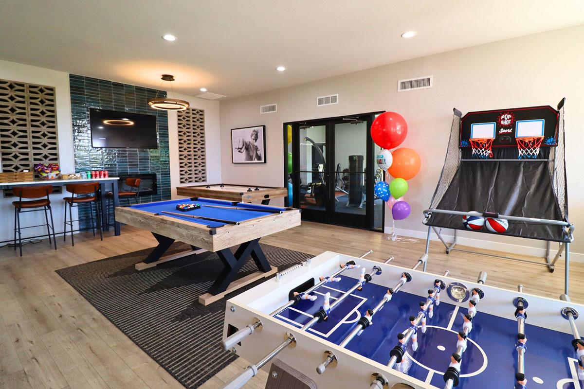 A recreational room featuring a blue pool table, a foosball table, and a mini basketball game. The space is decorated with colorful balloons and has a bar area with high stools. Large windows provide natural light, enhancing the inviting atmosphere for leisure activities.