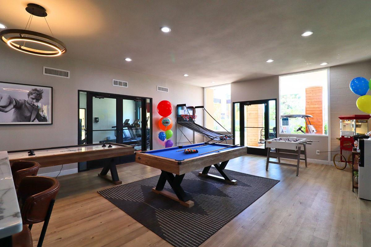 Interior space featuring a game room with a billiards table and a foosball table. There are colorful balloons and a popcorn machine in the corner. Large windows provide natural light, and workout equipment is visible in the background. The decor is modern, creating a vibrant and inviting atmosphere.