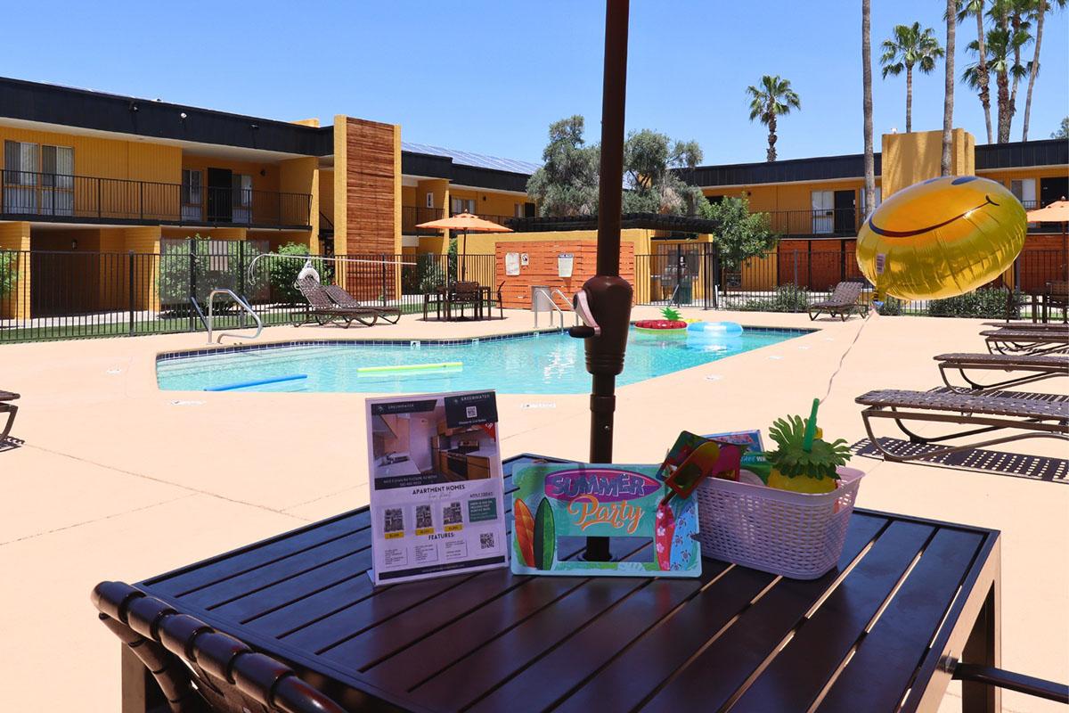 A sunny pool area surrounded by lounge chairs and palm trees. In the foreground, there's a table with a "Summer Party" decoration, a brochure, and a yellow balloon. The pool is inviting, with umbrellas providing shade in the background and bright blue skies overhead.