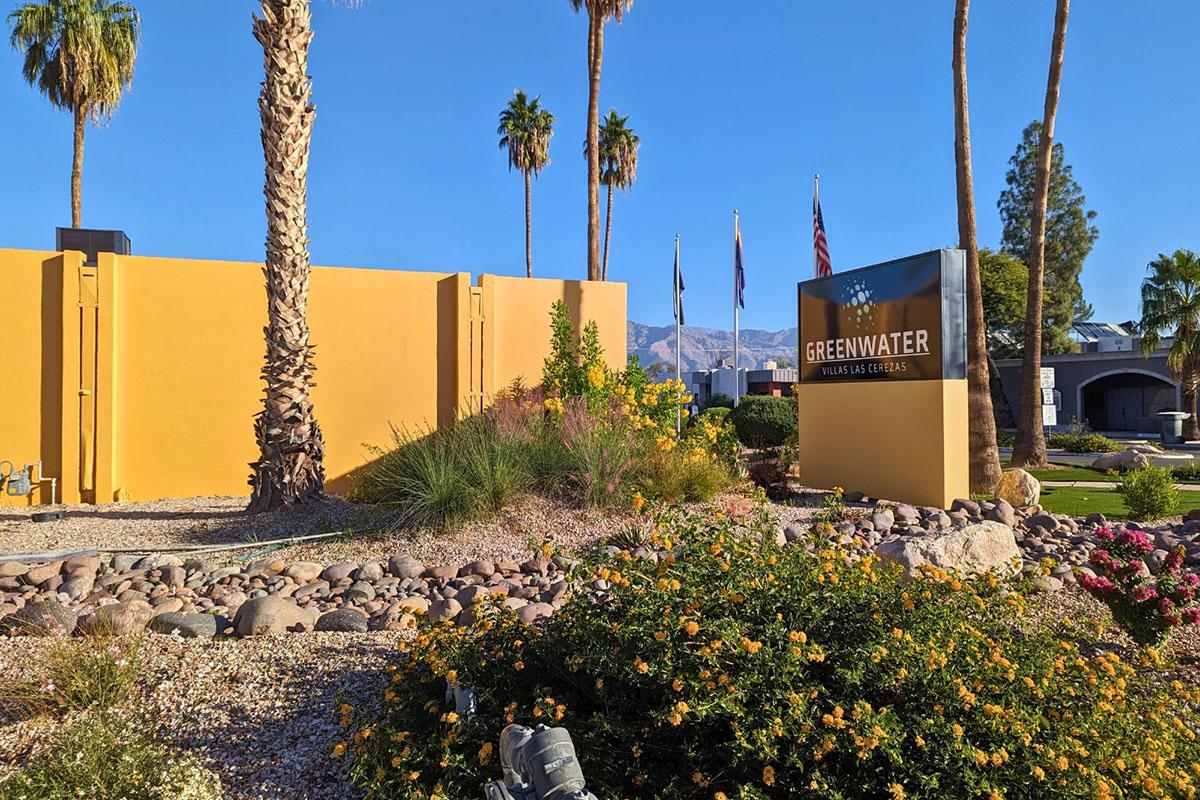 A colorful landscape featuring a prominent sign that reads "GREENWATER." The area is adorned with vibrant flowers and shrubs, framed by palm trees. In the background, a clear blue sky and distant mountains are visible, contributing to a sunny, inviting atmosphere.