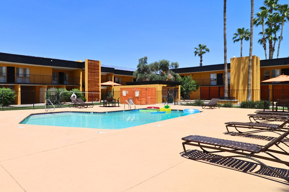 A sunny outdoor pool area featuring a clear blue swimming pool surrounded by lounge chairs. Palm trees and tropical landscaping enhance the setting, with an apartment building in the background. Umbrellas provide shade for relaxation, and pool floats are scattered on the water.