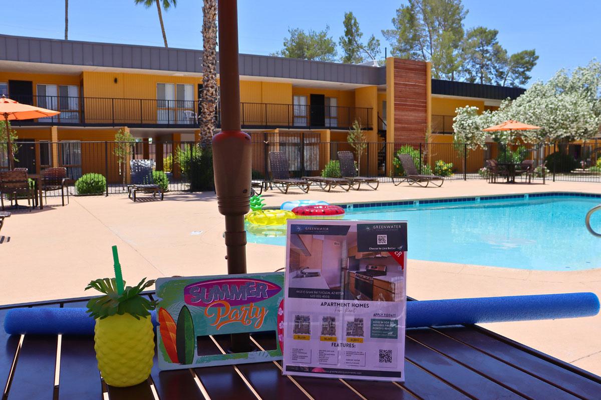 A vibrant pool area featuring lounge chairs, umbrellas, and tropical-themed decorations. In the foreground, there's a colorful "Summer Party" sign and an advertisement for apartment homes, set against the backdrop of a clear blue pool and sunny weather. Lush greenery adds to the relaxing atmosphere.