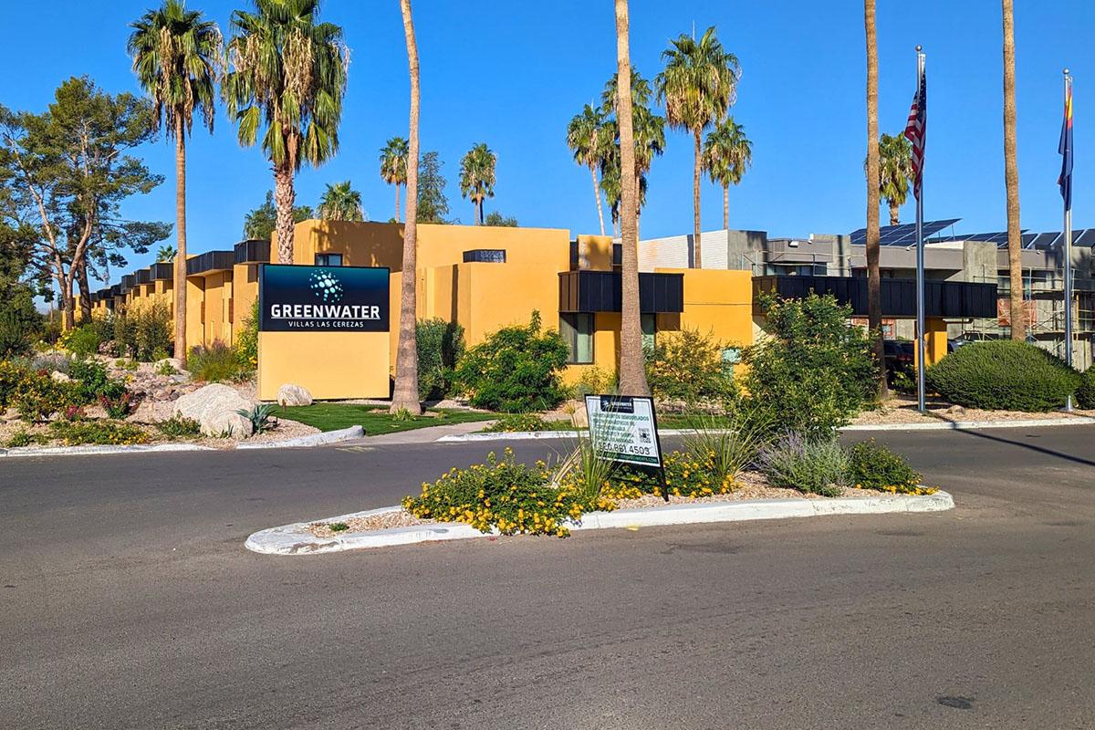 View of Greenwater Apartments, a modern residential complex, featuring yellow and black architecture surrounded by palm trees and landscaped gardens. A sign displays the apartment name and the entrance to the community, with a clear blue sky above.