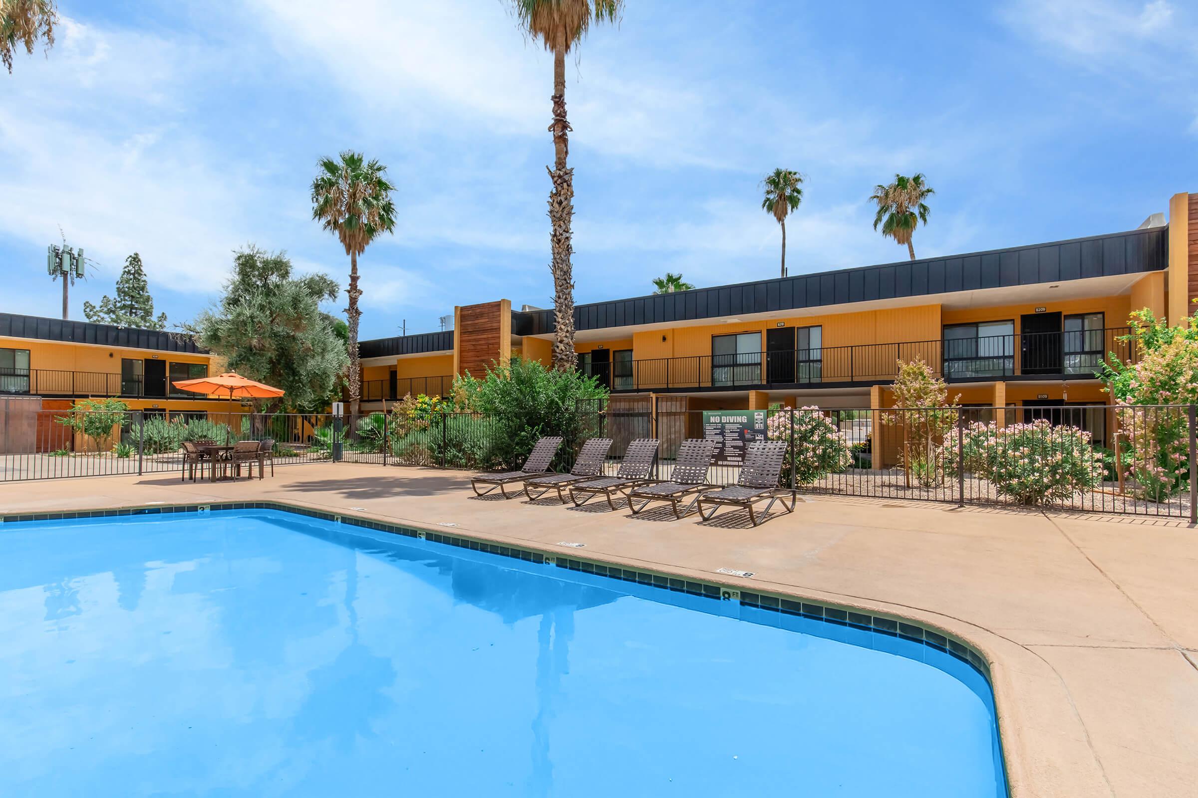 A refreshing outdoor pool area with several lounge chairs positioned around it, surrounded by palm trees and greenery. In the background, a two-story building painted in yellow and black features balconies. A large orange umbrella provides shade over a seating area. Clear blue sky enhances the vibrant scene.