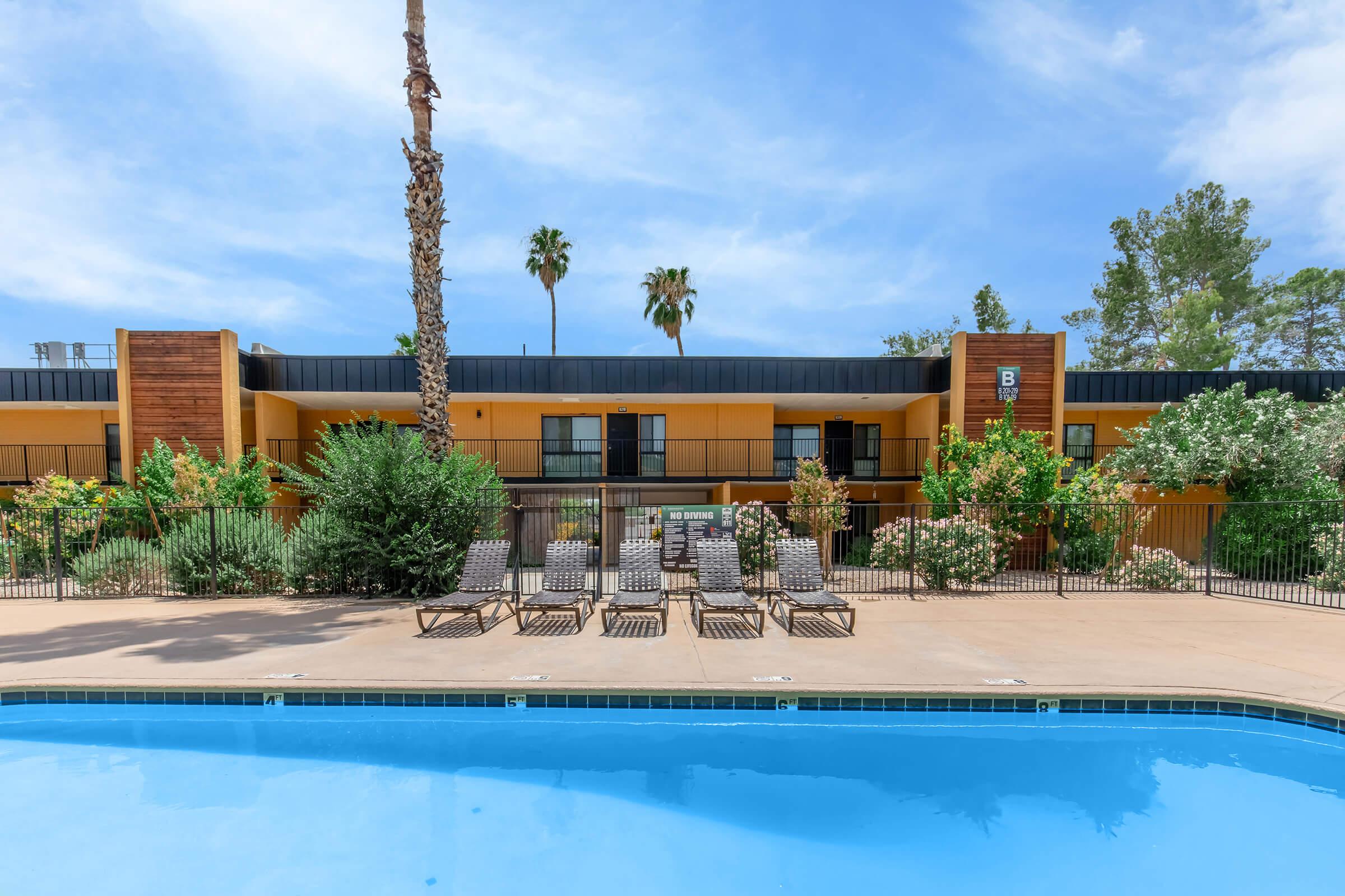 A view of a swimming pool surrounded by lounge chairs and lush greenery. In the background, there is a two-story building with a warm yellow facade and black accents, framed by palm trees under a clear blue sky. The setting conveys a relaxed, inviting atmosphere.