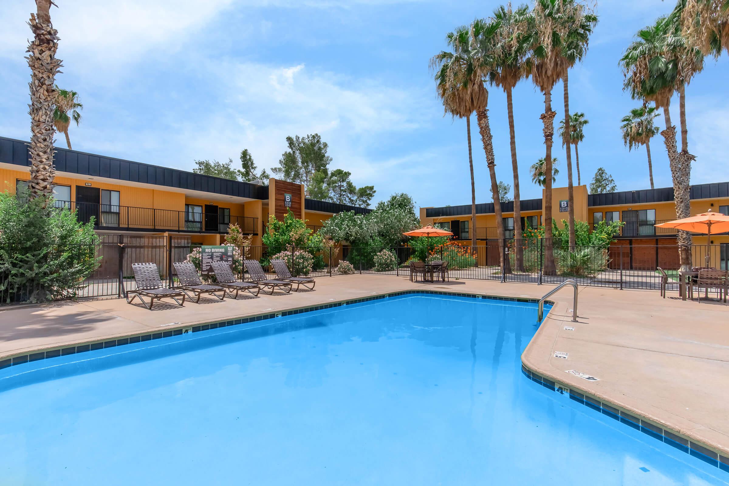 A clear blue swimming pool surrounded by lounge chairs and palm trees. Orange umbrellas provide shade near the pool area. In the background, a building with a landscaped area is visible under a clear blue sky.