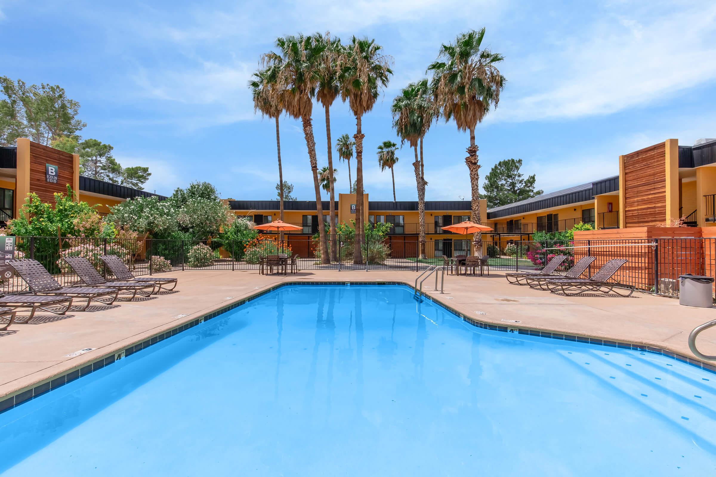 A clear blue swimming pool surrounded by lounge chairs and palm trees, with bright orange umbrellas providing shade. The background features a modern hotel facade with greenery and a sunny sky.