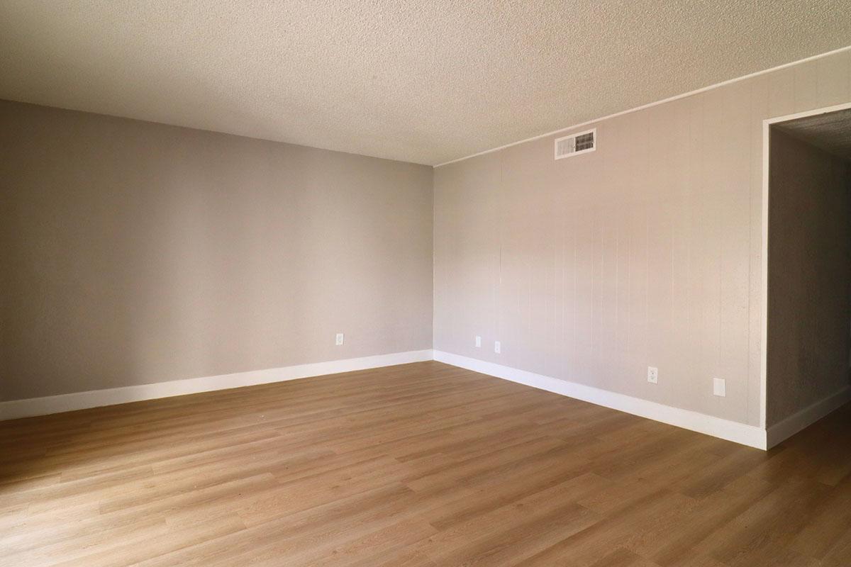 A spacious, empty room with light gray walls and wooden flooring. One corner has a wall with a small vent, and there is a doorway leading to another room visible on the right side. Natural light fills the space, creating a bright and inviting atmosphere.