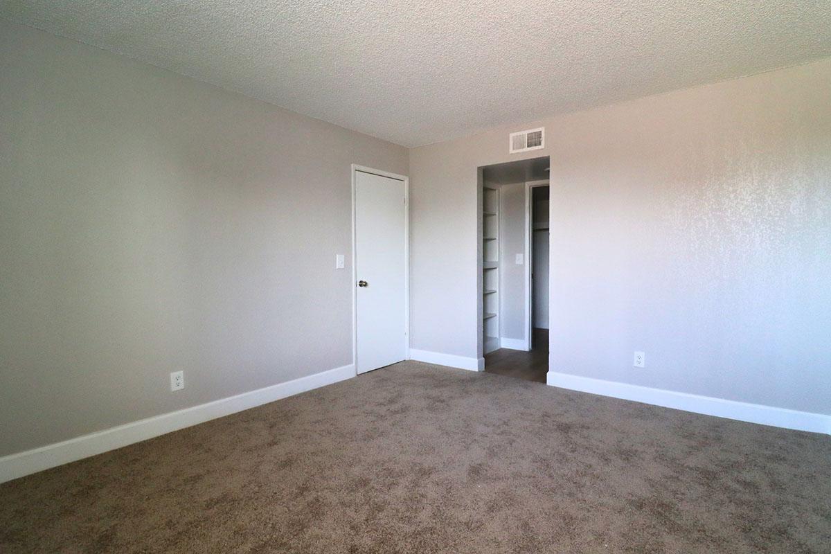 Empty room with light gray walls and beige carpet. There is a closed white door on the left and a small open closet or storage space on the right with built-in shelves. The space is well-lit and minimalist, creating a clean and neutral environment.