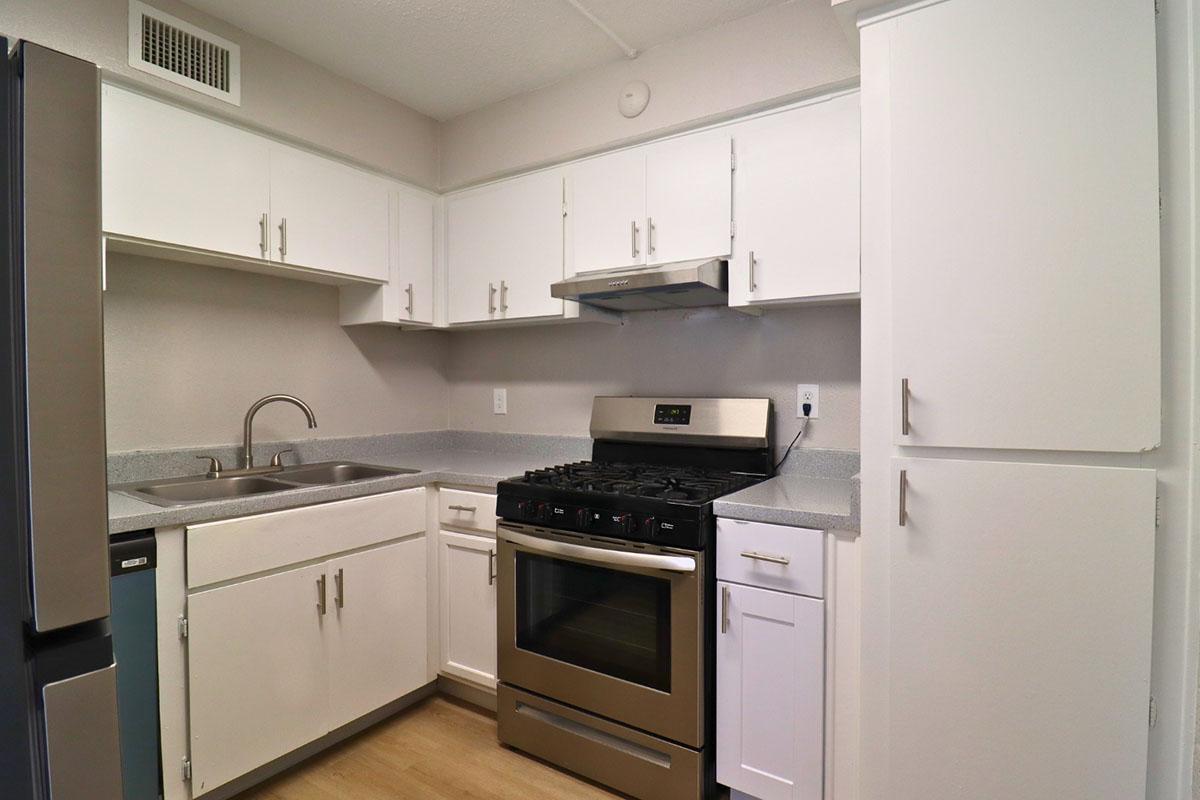 A clean, modern kitchen featuring white cabinets, a stainless steel gas stove, a stainless steel sink with a single faucet, and gray countertops. The space has a light and airy feel, with natural light coming in and wood-style flooring.