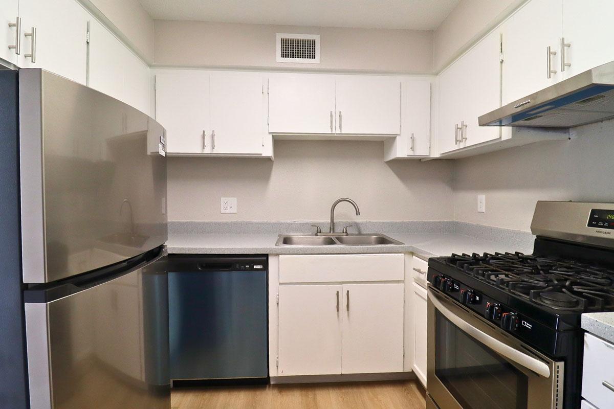 Modern kitchen featuring a stainless steel refrigerator, dishwasher, and gas stove. The space has white cabinetry and a gray countertop, with a double sink positioned between the appliances. The flooring is light-colored, enhancing the clean and bright atmosphere of the kitchen.