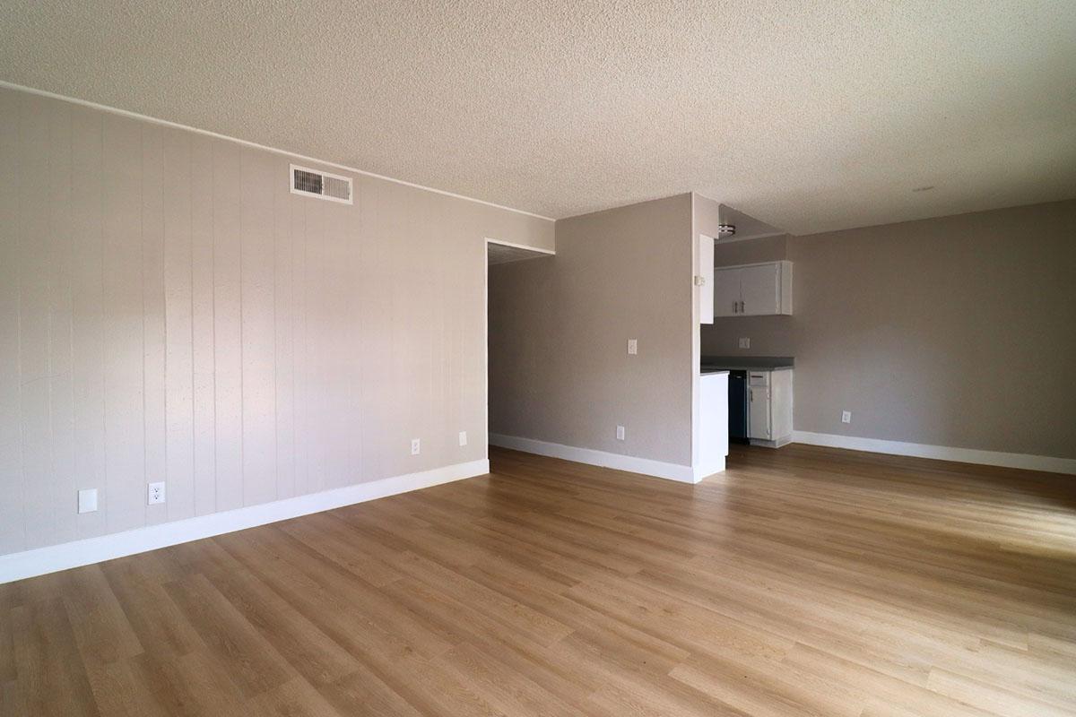 A spacious, empty room with light-colored walls and wooden flooring. On the left, there's a partially visible wall with a vent, and to the right, a small kitchen area with white cabinetry and a countertop. Natural light shines through, enhancing the airy feel of the space.