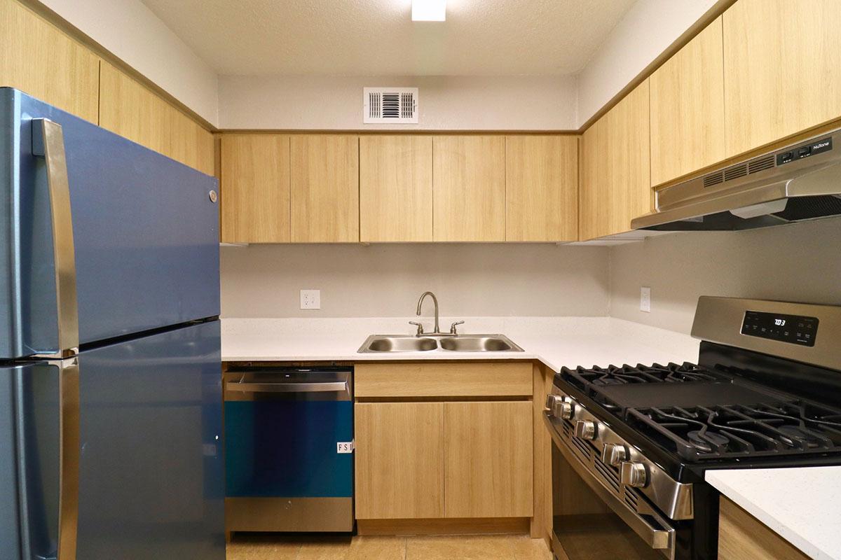 A modern kitchen featuring light wooden cabinetry, a stainless steel refrigerator, a stainless steel dishwasher, a double sink, and a gas stove. The countertops are white, and the flooring is beige tile. The space is well-lit with an overhead light fixture.