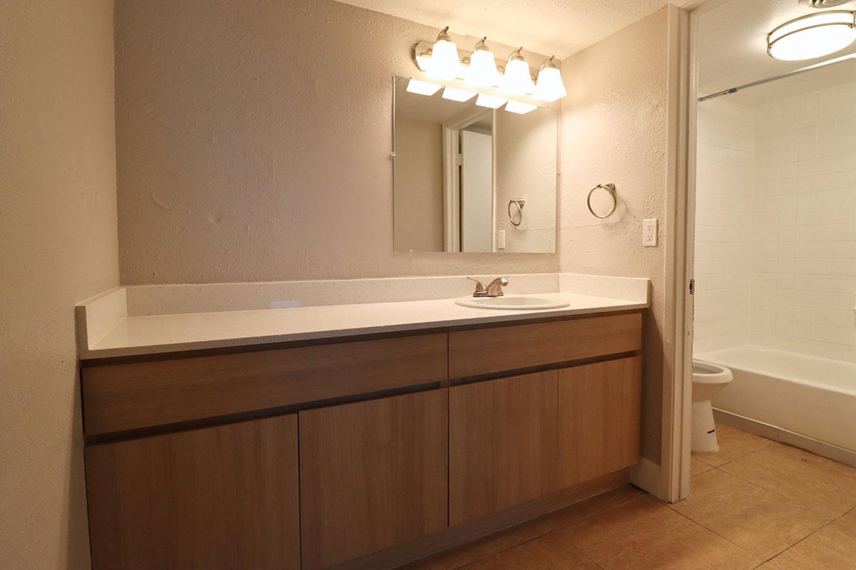 A simple modern bathroom featuring a wooden vanity with a white countertop and sink. Above the sink, there is a rectangular mirror and three light fixtures. To the right, a shower area is visible with a white bathtub and tiled wall. The floor is tiled with a warm brown color, creating a clean, neutral aesthetic.