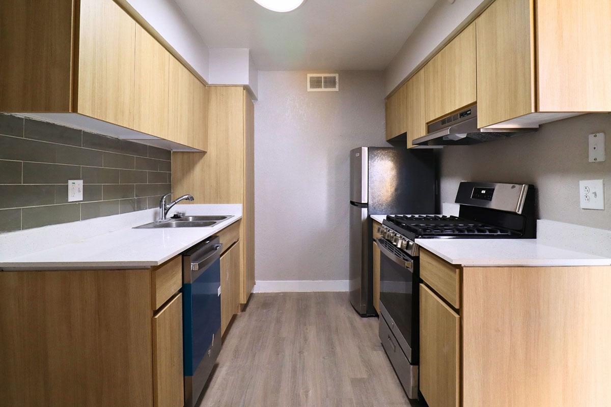 Modern kitchen featuring light wood cabinetry, a stainless steel refrigerator, and a gas stove. The countertop is white with a gray tiled backsplash. The space is well-lit, with a clean and contemporary design, ideal for cooking and meal preparation.