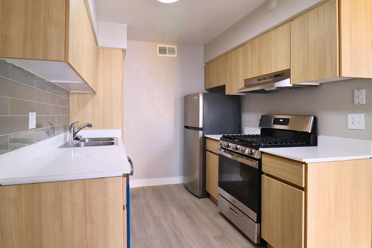 Modern kitchen featuring light wood cabinetry, a stainless steel refrigerator, and a gas stove with a hood. The countertop is white with a subtle texture, and there's a single-basin sink. Gray tile backsplash complements the light color scheme. The floors are light-colored wood laminate, creating a bright and contemporary atmosphere.