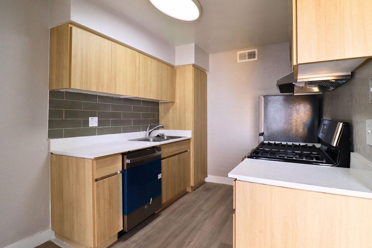 Modern kitchen featuring light wood cabinetry, a stainless steel sink, a gas stove, and a dishwasher. The countertops are white with a subtle texture, and there’s a gray tile backsplash. The flooring is a light-colored laminate, creating a bright and inviting space.