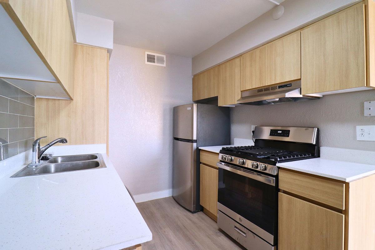 A modern kitchen featuring light wood cabinetry, a stainless steel refrigerator, a gas oven with a range hood, and a spacious countertop. The back wall is tiled in a neutral color, and there is a double sink on the left with a sleek faucet. Bright, minimalistic design creates an inviting atmosphere.