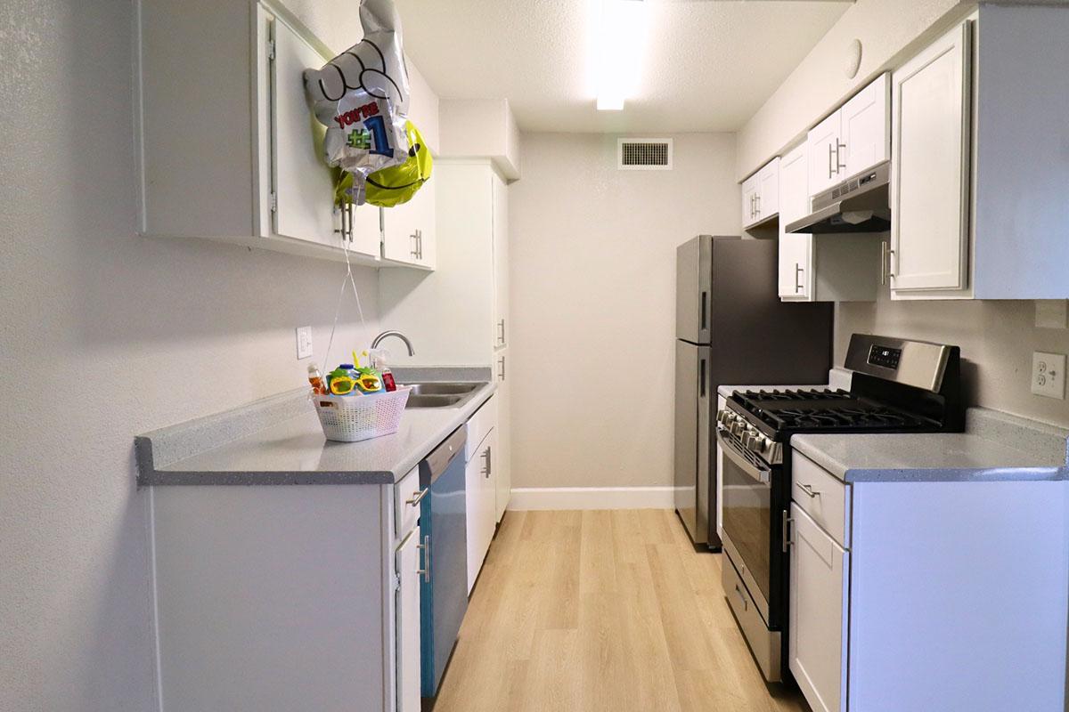 A modern kitchen featuring white cabinets, a stainless steel refrigerator, and a gas stove. The countertops are light gray, and there is a basket on the counter with colorful items. A helium balloon is attached to the cabinet, adding a festive touch to the space. Bright lighting enhances the airy atmosphere.