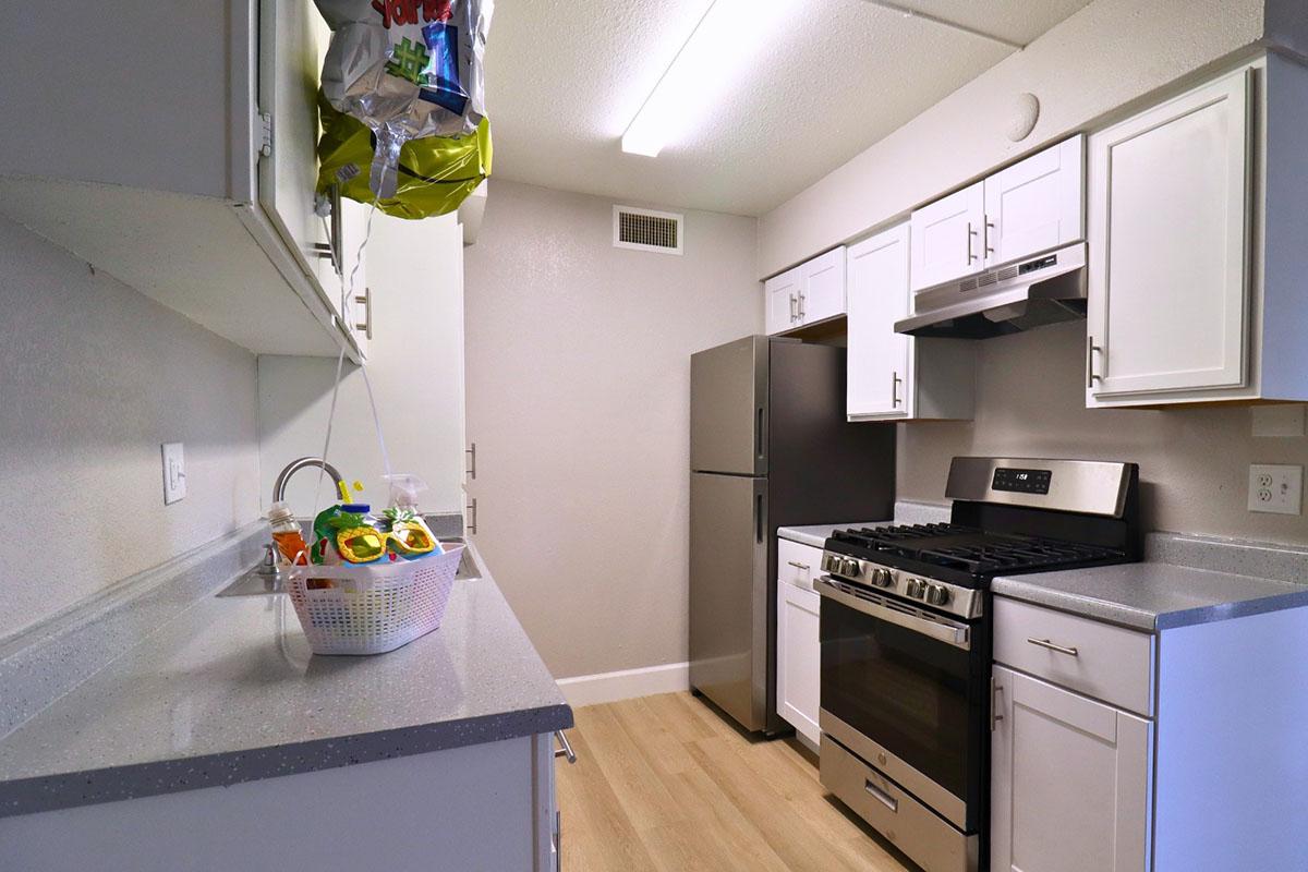 A modern kitchen featuring white cabinetry, stainless steel appliances, and a gray countertop. A silver refrigerator and a black gas stove are visible. A basket filled with colorful kitchen items sits on the countertop, and a few balloons are tied to a cabinet handle, adding a festive touch.