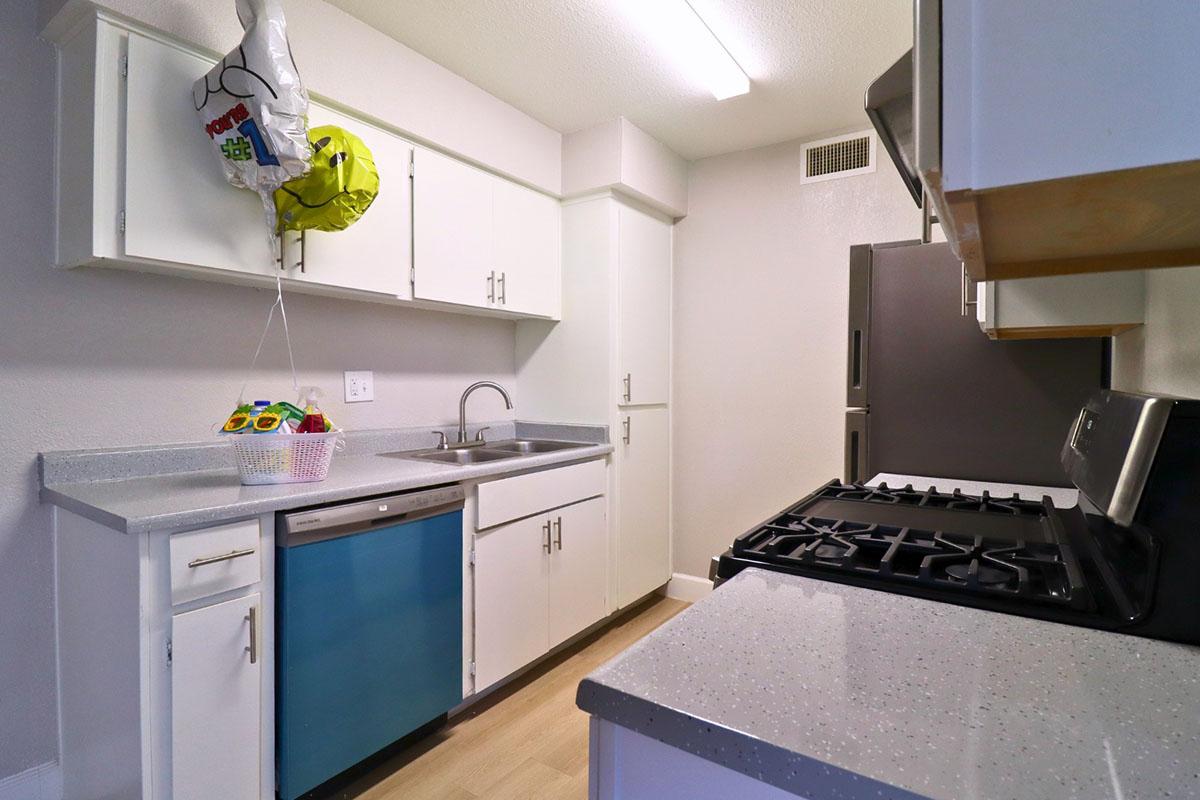 A modern kitchen featuring white cabinetry, a stainless steel oven, and a blue refrigerator. The countertop is light gray with a sink, and there is a small basket with colorful items. A balloon is floating near the ceiling, adding a festive touch to the space. The flooring is light wood.