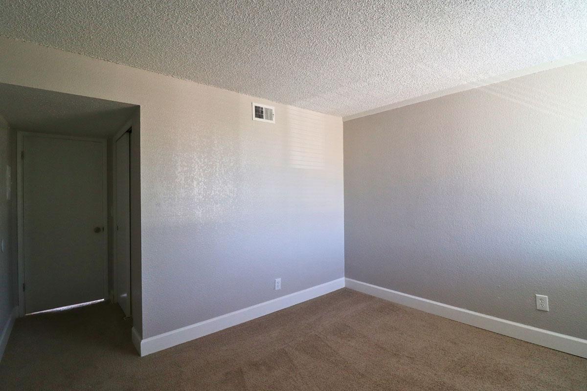 A vacant room with light gray walls and a tan carpet. There is a closed door on the left leading to another space, and a small window on the far wall allowing natural light to enter. The room appears clean and empty, suitable for decoration or furniture placement.