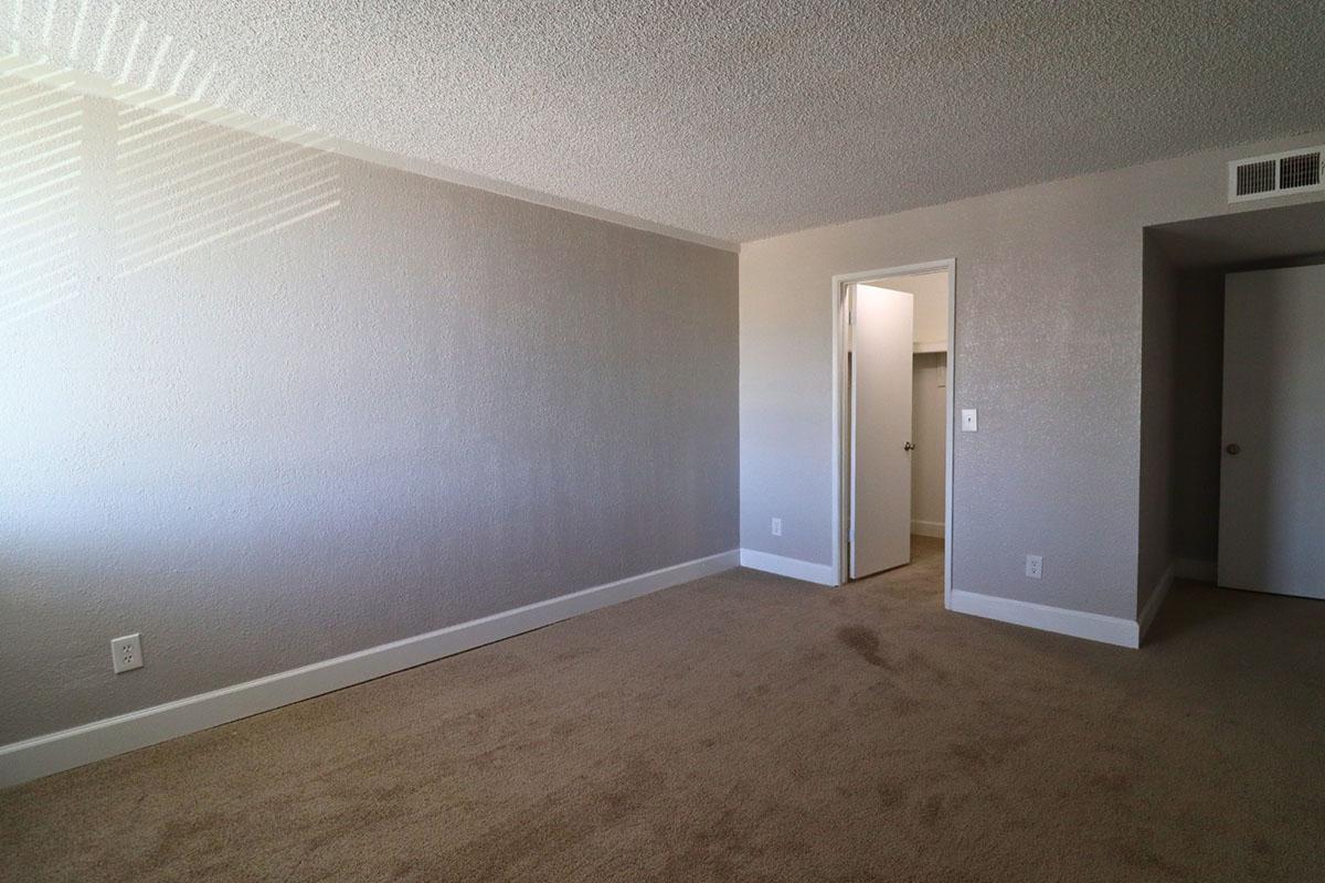 A vacant room with light gray walls and carpet. The space features a door on the left leading to a closet, and another door on the right leading outside of the room. Sunlight casts shadows on the wall, creating a striped pattern. The overall atmosphere is calm and uncluttered.