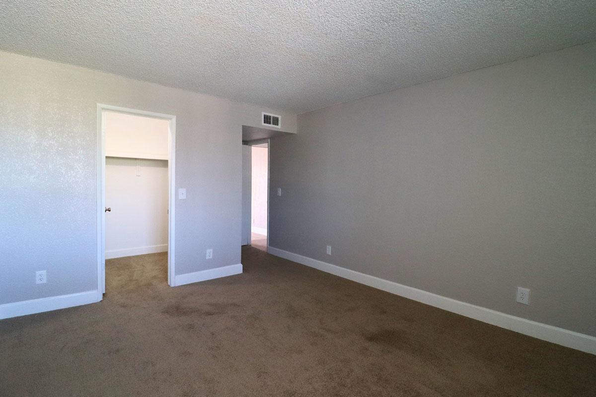 A spacious, empty room with light beige carpet and light gray walls. A doorway on the left leads to a small closet, while an open door in the background shows another area. Natural light is coming in, giving a bright and airy feel to the space.