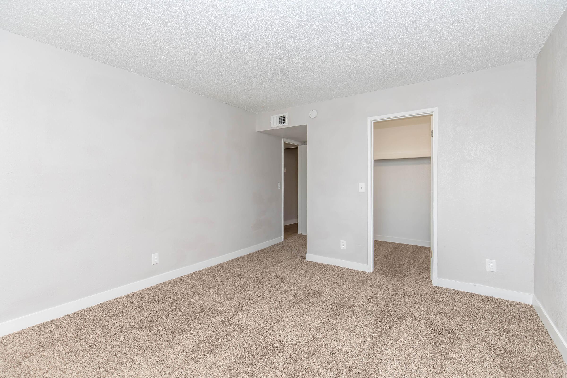 A vacant room with light gray walls and beige carpet. There is a doorway leading to a closet on the right and a wall with a slight indentation for a potential entryway. The overall ambiance is bright and neutral, ideal for customization or furnishing.