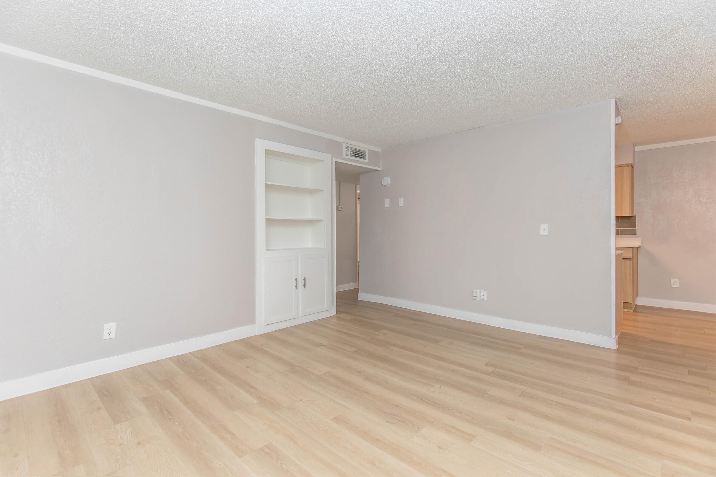 A spacious, unfurnished room with light gray walls and a light wooden floor. There is a built-in white shelving unit on the left side, and an open entryway leading to another room in the background. The overall atmosphere is bright and clean, with natural light illuminating the space.