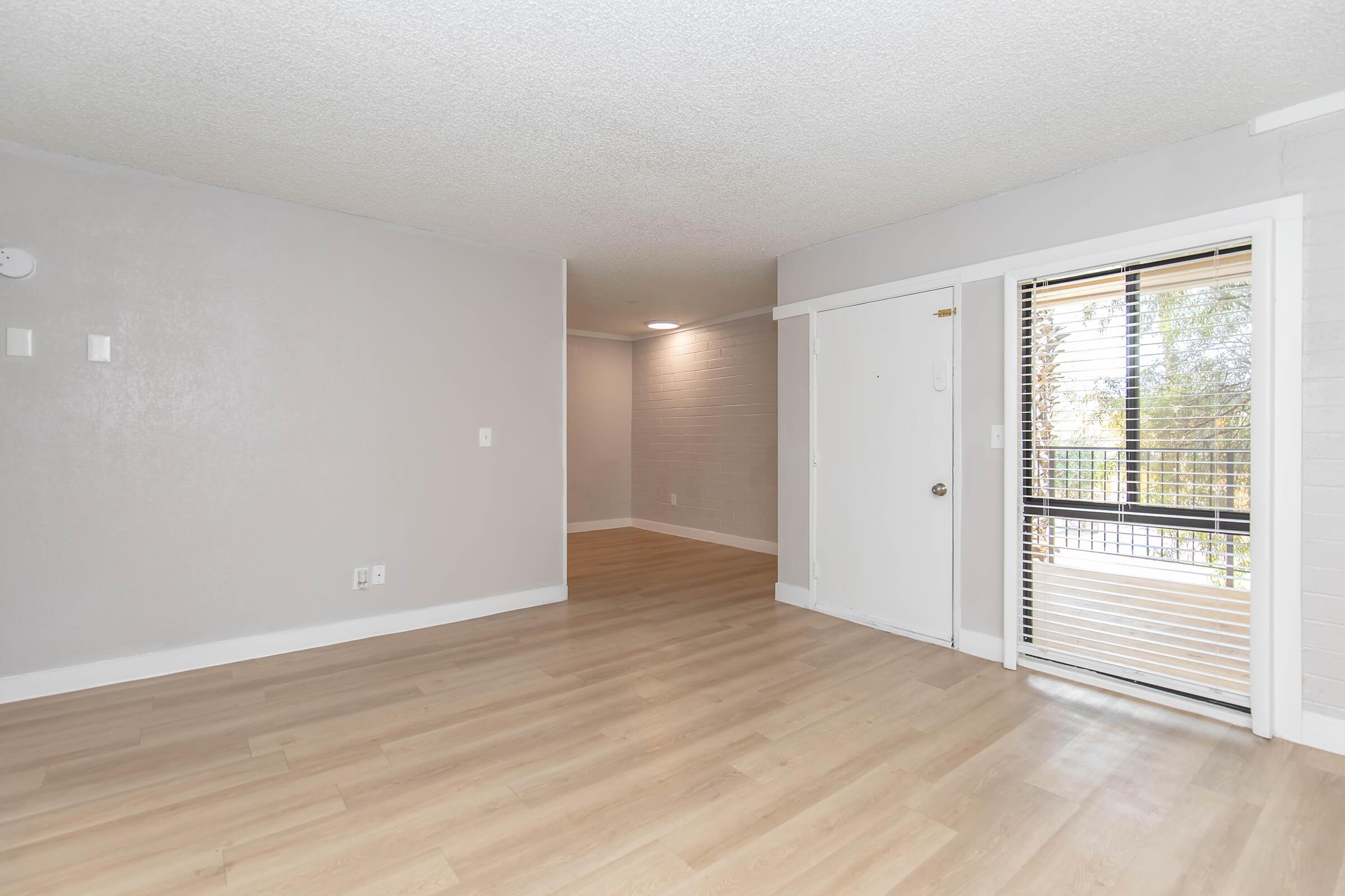 A spacious interior view of a modern living area with light-colored wooden floors. The walls are painted in soft gray tones, and there is a large window with blinds that lets in natural light. The layout appears open, leading to another room off to the side.