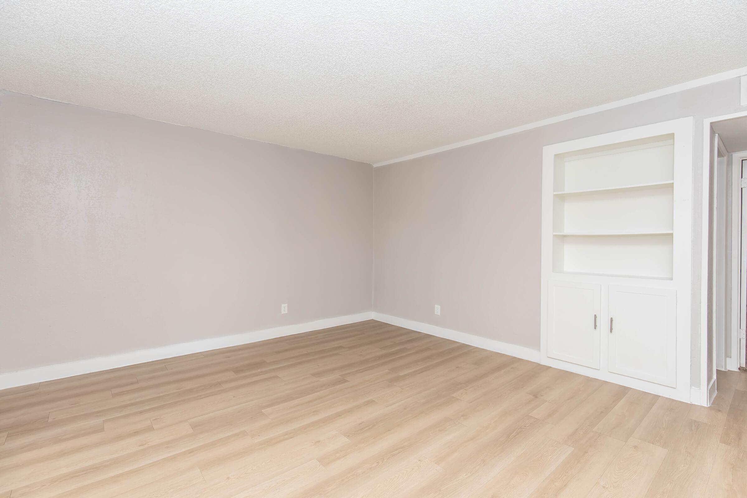 An empty room with light gray walls and hardwood flooring. There is a built-in white shelving unit on one side, while the rest of the space is unobstructed, creating a clean and minimalist appearance. Natural light may be coming in, highlighting the simplicity of the design.