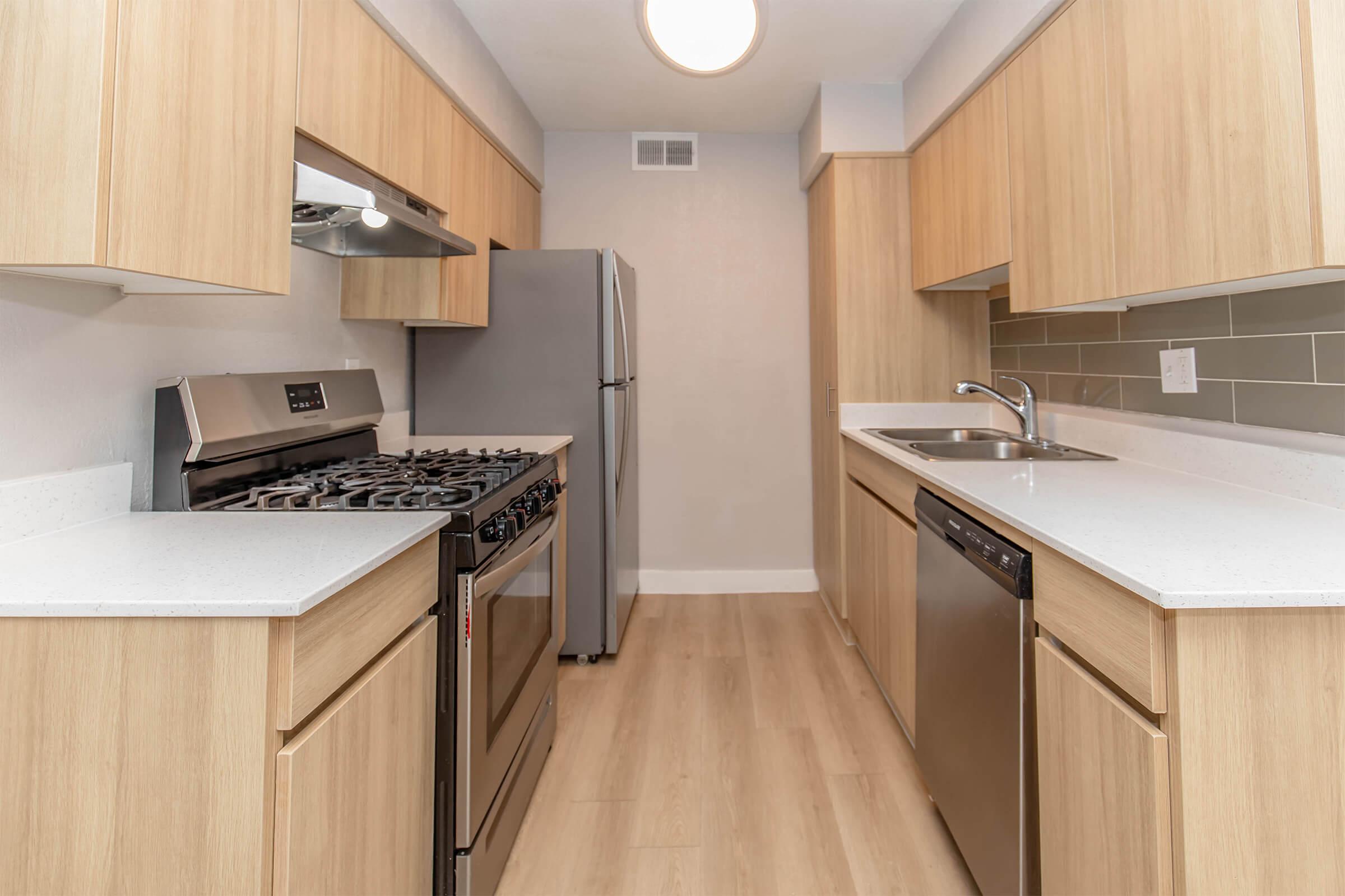Modern kitchen with wooden cabinetry, stainless steel appliances including a gas stove and refrigerator. The countertops are white, complemented by a sleek backsplash. The kitchen features ample cabinet space and a sink with a faucet, set against a neutral wall color and light-colored flooring.