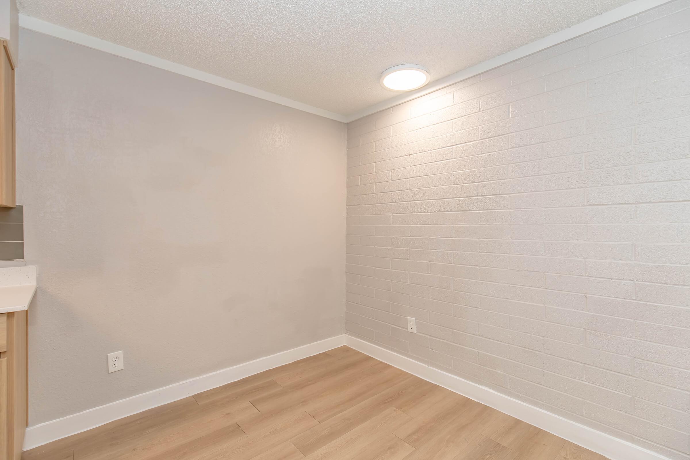 A well-lit, empty room with light gray painted walls and light wood flooring. The space features a ceiling light and is devoid of furniture or decorations, creating a clean and minimalist appearance.