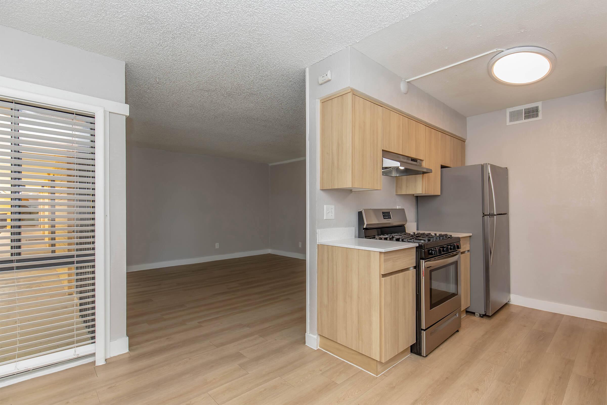 Modern kitchen with light wood cabinetry, stainless steel gas stove, and refrigerator. The room features a light-colored wall and flooring, with a large window allowing natural light. An open area adjacent for dining or living space, enhancing the spacious feel of the design.