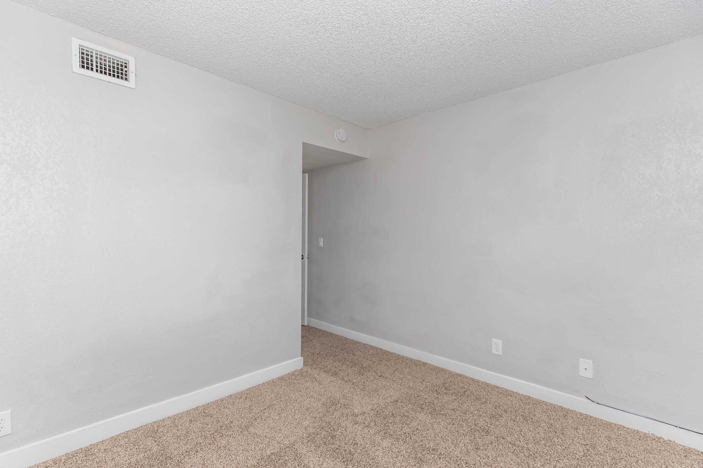 Empty room with a textured light gray wall and a beige carpeted floor. There is an open doorway leading to another space, and a small vent on the wall. The overall atmosphere is neutral and minimalistic.