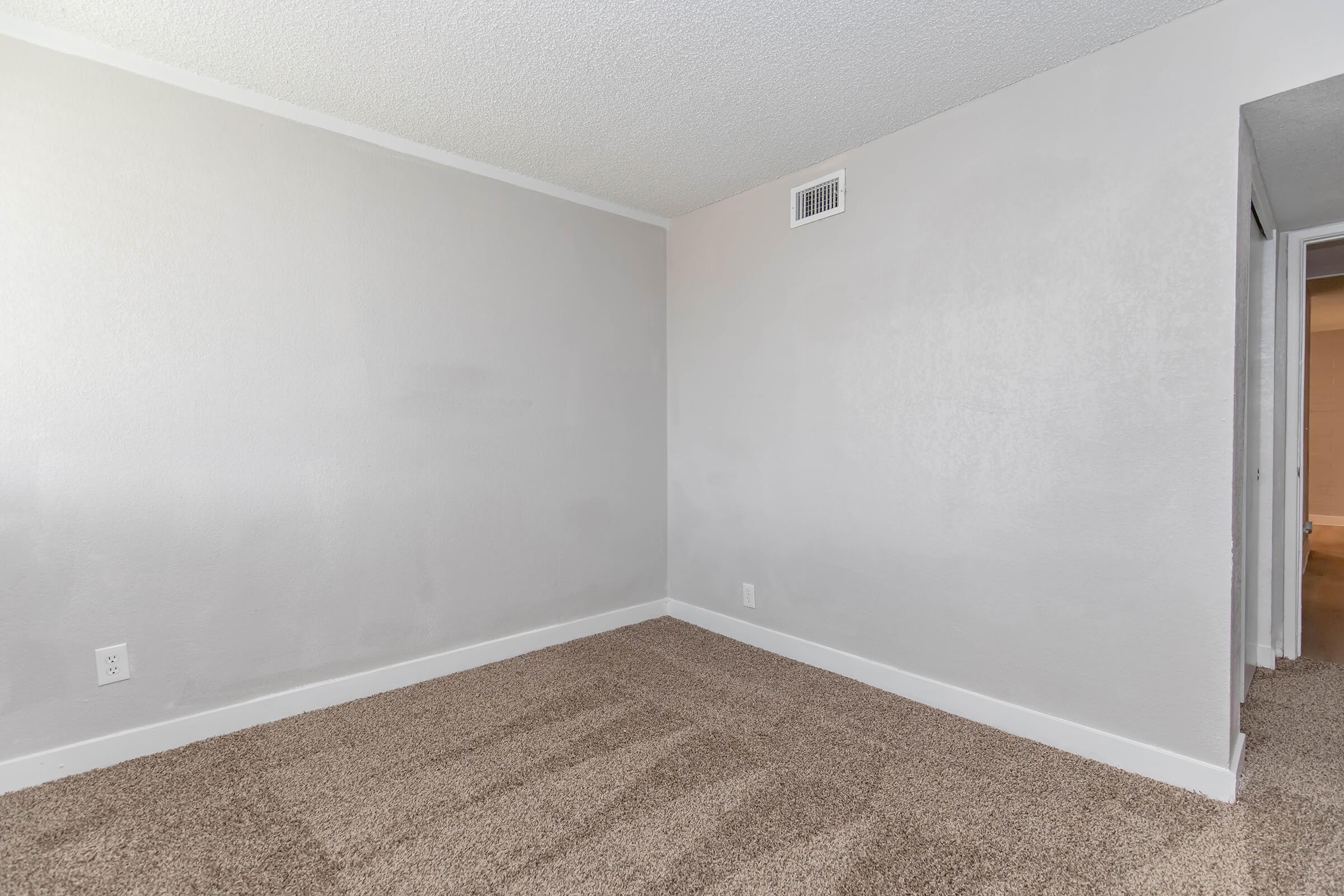A lightly colored empty room with a textured wall and plush brown carpet. The room is well-lit and features a small vent on the wall. There are doorways leading to adjacent spaces, but no furniture or decorations present. The overall atmosphere is clean and minimalistic.