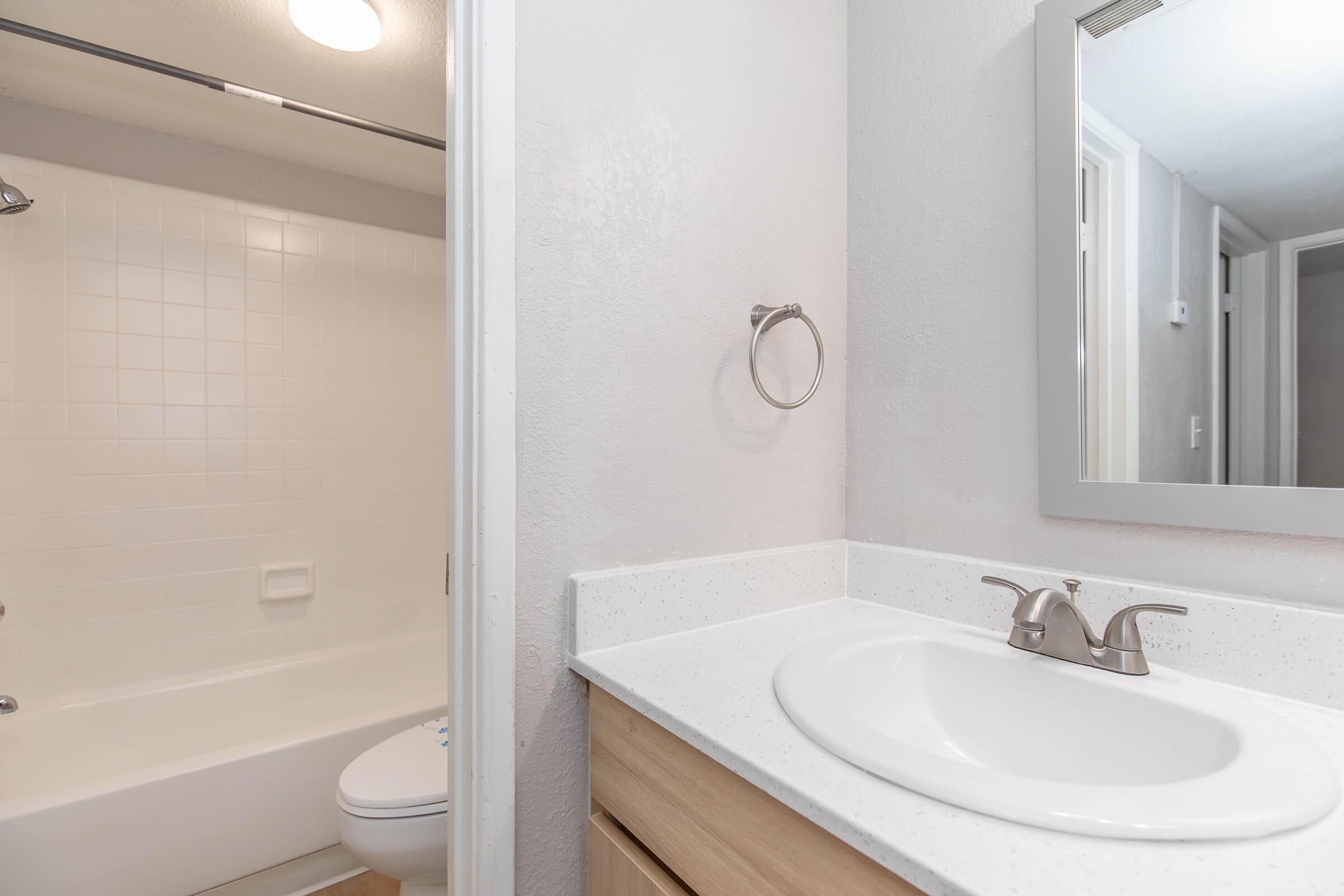 A clean and modern bathroom featuring a white countertop with a sink and faucet, a towel ring, and a large mirror. In the background, there is a shower/tub combination with white tiles and a light fixture above. The walls are painted in a soft neutral color, creating a bright and airy atmosphere.